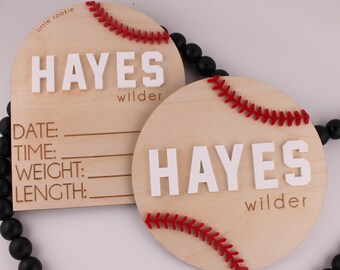 May include: Two wooden baseball-themed baby announcement signs. One sign is a half-circle with the text "little rookie" and "HAYES wilder". The other sign is a circle with the text "HAYES wilder" and a red baseball stitch design.