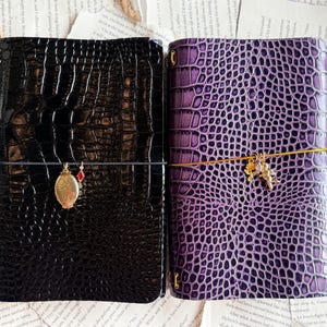 May include: Two leather journals with a crocodile skin texture. One is black, the other is purple. Each journal has a decorative charm and an elastic closure. The journals are lying on a background of vintage paper and stamps.