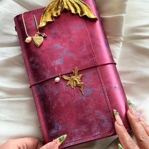 May include: A rectangular journal with a distressed deep red and purple cover, featuring a gold-toned mermaid tail bookmark at the top, a gold heart charm on a wire clip, and a gold fairy charm attached to a thin red elastic band across the centre.