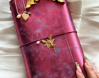 May include: A rectangular journal with a distressed deep red and purple cover, featuring a gold-toned mermaid tail bookmark at the top, a gold heart charm on a wire clip, and a gold fairy charm attached to a thin red elastic band across the center.