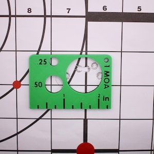 May include: A bright green sight alignment tool with circular cutouts and inch measurements. The tool has the text "1 MOA" and "2 in" printed on it. The tool is placed on a white target with black grid lines and red circles.