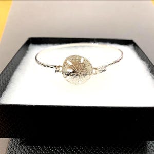 Op de afbeelding: Een zilverkleurige armband met een zanddollar-bedel. De armband wordt gepresenteerd in een zwarte doos met een witte binnenkant. De zanddollar-bedel is rond met gedetailleerde patronen.