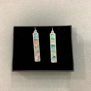 May include: A pair of rectangular dangle earrings with iridescent abalone shell inserts set in silver-toned metal. The earrings are displayed in a black jewelry box against a gray background. The abalone shell displays shades of green, blue, pink, and purple.
