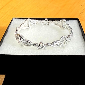 Könnte beinhalten: Ein silberfarbenes Armband mit einem komplizierten keltischen Knotenmuster. Das Armband wird in einer schwarzen Schachtel mit weißem Innenraum präsentiert, die seine detaillierte Handwerkskunst zeigt. Das Design weist ineinander verschlungene Muster und symmetrische Formen auf.