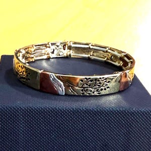 Puede incluir: Una pulsera elástica con eslabones metálicos rectangulares alternos en tonos oro, cobre y plata. Los eslabones están decorados con motivos de ramas de árboles grabados y paneles lisos de colores sólidos, creando un aspecto metálico multicolor.