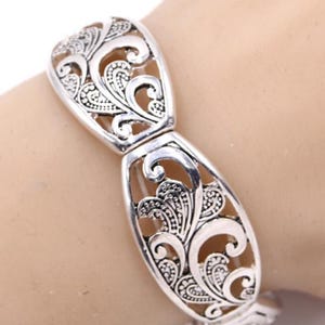 以下が含まれることがあります： シルバーカラーのブレスレット、透かし彫りデザイン。ブレスレットは、渦巻く葉と花の要素の装飾的で対称的なパターンが特徴です。ブレスレットは金属製で、留め金が付いています。