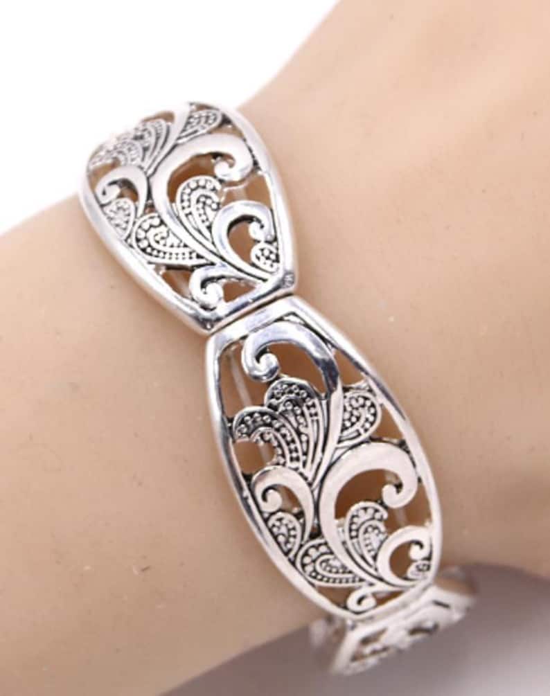 以下が含まれることがあります： シルバーカラーのブレスレット、透かし彫りデザイン。ブレスレットは、渦巻く葉と花の要素の装飾的で対称的なパターンが特徴です。ブレスレットは金属製で、留め金が付いています。