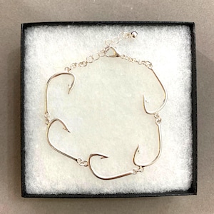 Puede incluir: Una pulsera de tono plateado que presenta cinco dijes de anzuelo conectados. La pulsera incluye un diseño de cadena fina con un cierre de mosquetón y una cadena extensora ajustable con una pequeña cuenta redonda en el extremo, dentro de una caja.