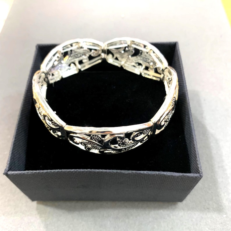 以下が含まれることがあります： 装飾的な透かし彫りのデザインが施されたシルバーカラーのブレスレット。ブレスレットは、それぞれ詳細な花柄とスクロール模様が特徴の複数の関節セグメントで構成されています。濃い灰色の箱の中の黒いベルベットの表面に置かれています。