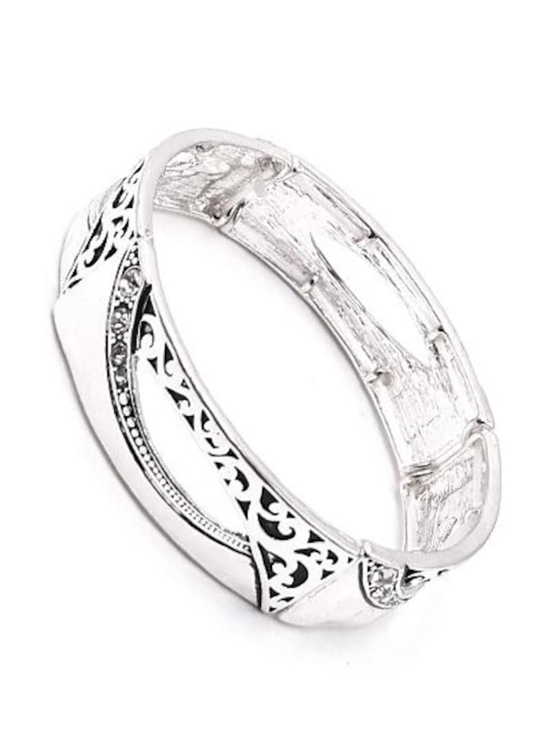 May include: Silver-toned hinged bracelet with a decorative design. The bracelet features a series of rectangular links, with intricate black cut-out patterns and small, clear gemstones. The bracelet is shown against a white background.
