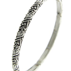 May include: A silver bangle bracelet with an intricate, ornate design. The bracelet is smooth and polished, with a delicate, detailed pattern.