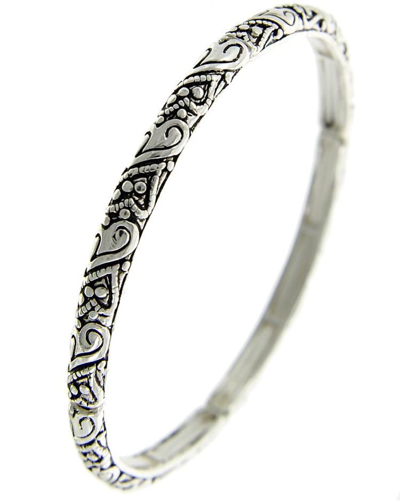 May include: A silver bangle bracelet with an intricate, ornate design. The bracelet is smooth and polished, with a delicate, detailed pattern.