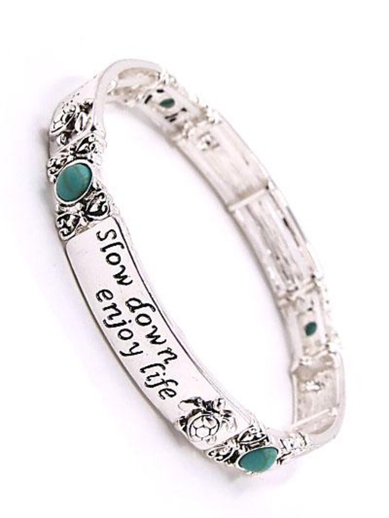 Peut inclure: Bracelet extensible argent&eacute; avec des accents turquoise et l'inscription "slow down enjoy life". Le bracelet pr&eacute;sente des motifs d&eacute;coratifs de tortues et de fleurs. Un accessoire &eacute;l&eacute;gant.