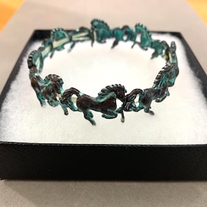 May include: A bronze-toned bracelet featuring a repeating pattern of horse figures. The horses have a verdigris patina, adding a touch of color. The bracelet is presented in a black box with a white interior, suggesting a jewelry item.