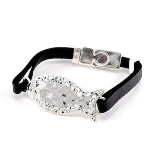 Puede incluir: Una pulsera de cuero negro con una pieza central plateada en forma de pez. El pez tiene un aspecto texturizado y martillado y está enmarcado por un borde plateado decorativo. La pulsera tiene un cierre plateado.