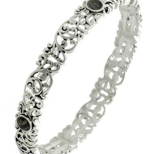 May include: A silver bangle bracelet with an openwork design. The bracelet features intricate scrollwork and two oval, dark grey stones. The metal has a polished finish, and the design is symmetrical.