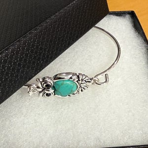 May include: A silver-toned metal bangle bracelet featuring an owl-shaped centerpiece. The owl has a smooth, oval-shaped turquoise stone as its body, with detailed metal wings and black circular eyes. The bracelet has a simple hook-style closure.