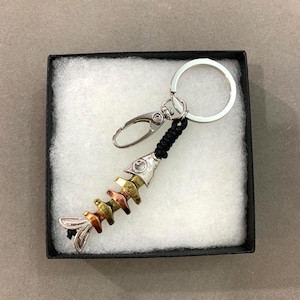 Puede incluir: Un llavero con forma de espina de pescado con una cabeza y cola de pez de metal plateado, y una serie de huesos de color cobre, latón y plata. El llavero incluye un mosquetón y un llavero. El llavero se presenta en una caja negra con acolchado blanco.