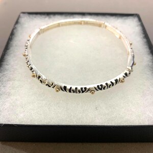 May include: A circular bangle bracelet featuring a silver-toned metal band with an intricate black engraved pattern and small, evenly spaced gold-toned spherical accents along the outer edge, presented inside a black jewelry box.