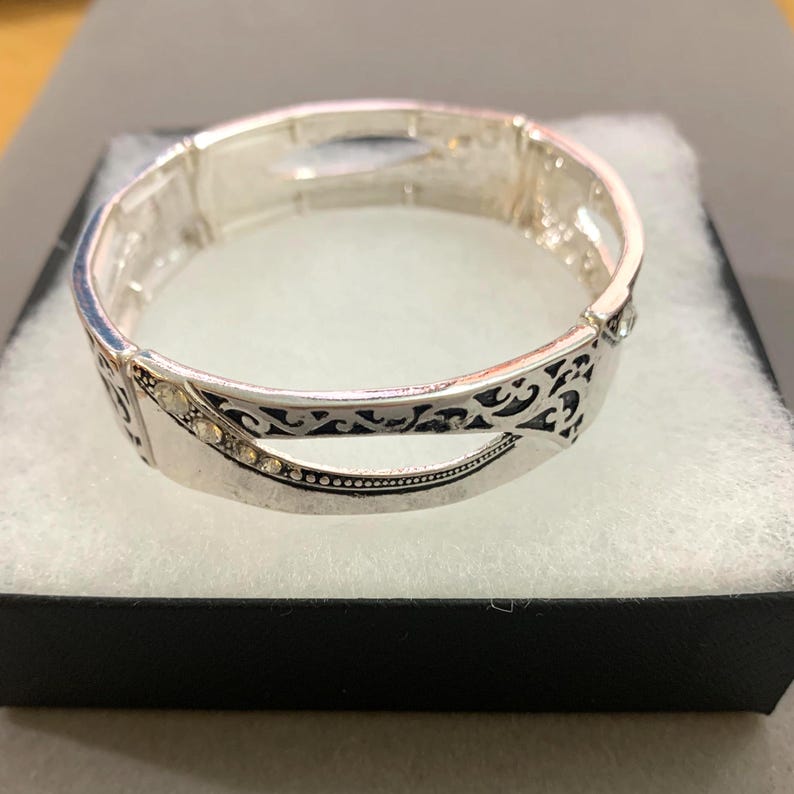 May include: Silver-toned bracelet with an ornate design. The bracelet features a series of small, clear gemstones and decorative cutouts. The bracelet is displayed on a white surface inside a black box.