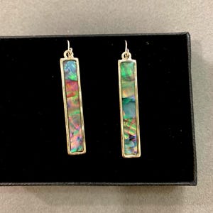 May include: A pair of rectangular dangle earrings with iridescent abalone shell inserts set in gold-colored metal frames. The earrings are displayed on a black surface.