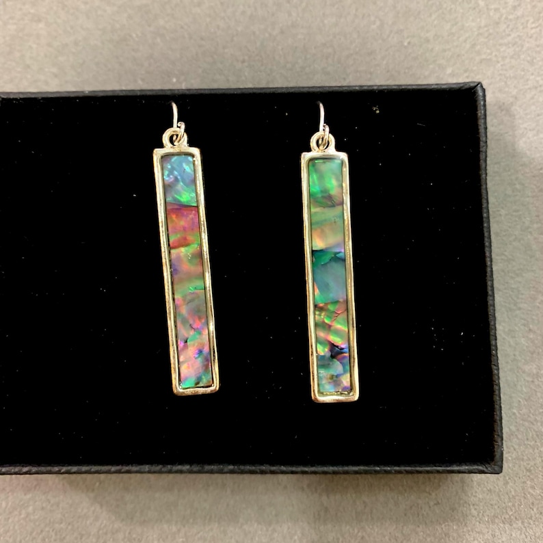 May include: A pair of rectangular dangle earrings with iridescent abalone shell inserts set in gold-colored metal frames. The earrings are displayed on a black surface.