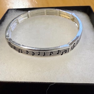 May include: Silver-coloured bracelet with a musical notes and staff design. The stretch bracelet is displayed in a black box with a white interior. The musical notes are black and the bracelet is approximately 18 cm in circumference.