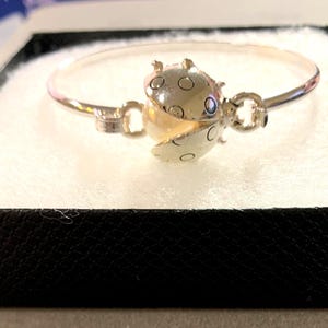 Pode incluir: Pulseira prateada com fecho de joaninha e uma pérola no centro. A joaninha tem pintas pretas e abre para revelar a pérola. A pulseira é apresentada numa caixa preta com interior branco.