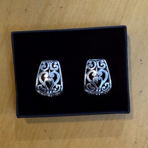 May include: A pair of silver-colored filigree earrings in a black box. The earrings have a rectangular shape with an openwork design featuring swirling patterns and a heart shape in the center. The earrings are displayed on a black velvet-like surface.