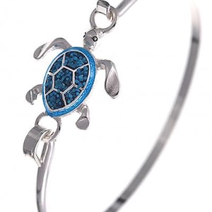 Può includere: Bracciale a cerchio argentato con un ciondolo a forma di tartaruga marina. La tartaruga ha un design a mosaico blu con un bordo argentato. Il bracciale è un design semplice ed elegante.