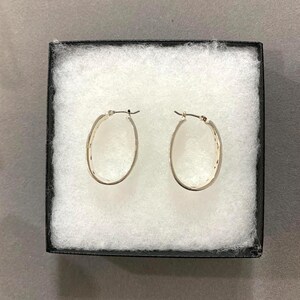 May include: A pair of oval-shaped silver hoop earrings displayed in a black box with white padding. The earrings have a textured design along the outer edge and a simple clasp closure. The earrings are a classic jewelry item.