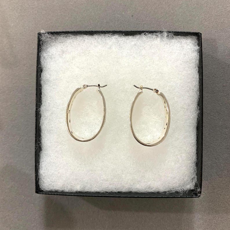 May include: A pair of oval-shaped silver hoop earrings displayed in a black box with white padding. The earrings have a textured design along the outer edge and a simple clasp closure. The earrings are a classic jewelry item.