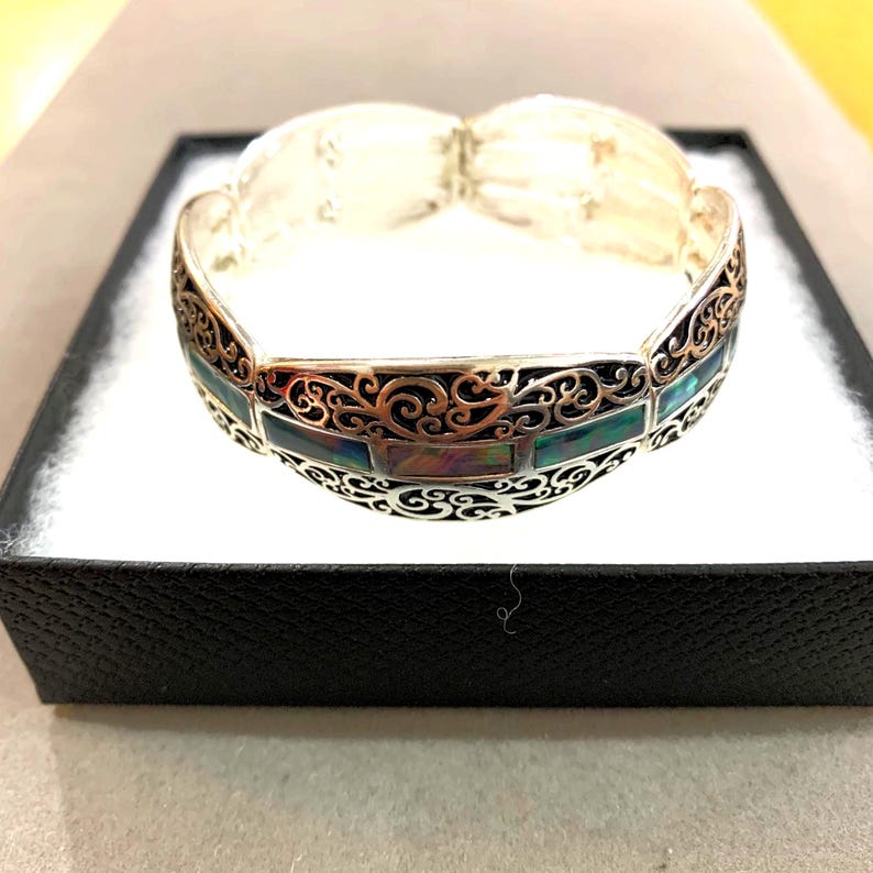 Pu&ograve; includere: Bracciale argentato con cerniera, con intricati filigrane e intarsi rettangolari iridescenti. Il bracciale &egrave; esposto in una scatola nera con interno bianco, che mette in risalto il suo design ornato e la sua maestria.