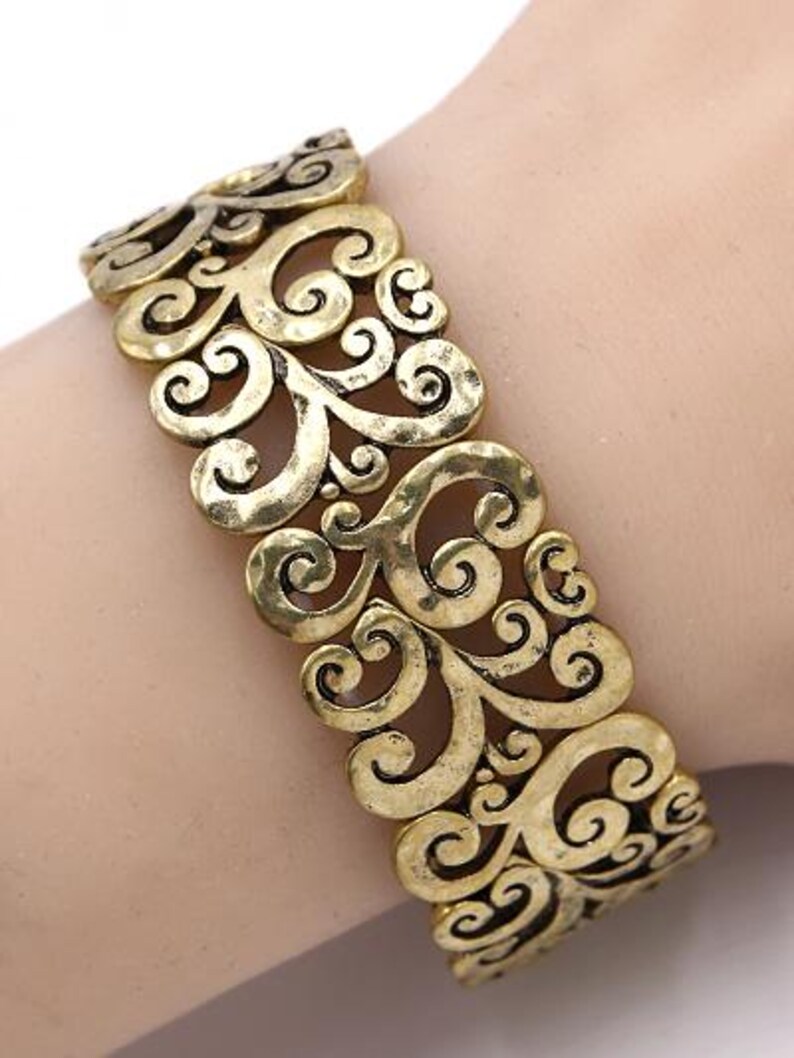 Może przedstawiać: Bransoletka z metalu w kolorze złotym z ozdobnym, skomplikowanym, ażurowym wzorem z wirujących, kwiatowych wzor&oacute;w.
