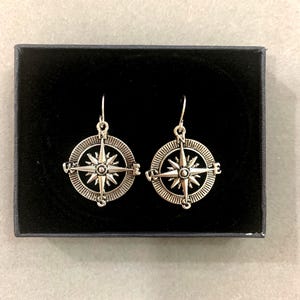 Puede incluir: Un par de pendientes de brújula plateados. Cada pendiente presenta un diseño de brújula detallado con una estrella central y marcadores direccionales. Los pendientes se muestran en una caja negra.