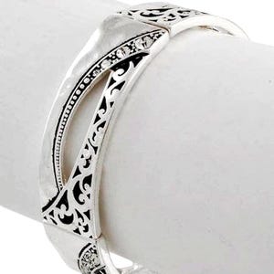 May include: Silver-toned hinged bracelet with intricate openwork and small, clear gemstones. The bracelet features a decorative design with a mix of smooth and textured surfaces. The bracelet is shown on a white background.