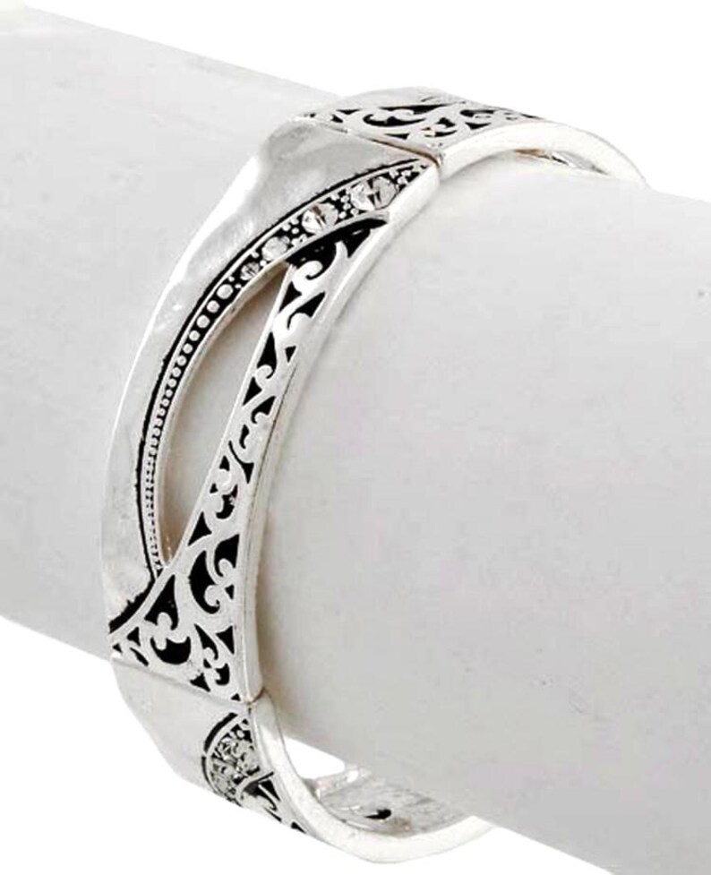 May include: Silver-toned hinged bracelet with intricate openwork and small, clear gemstones. The bracelet features a decorative design with a mix of smooth and textured surfaces. The bracelet is shown on a white background.