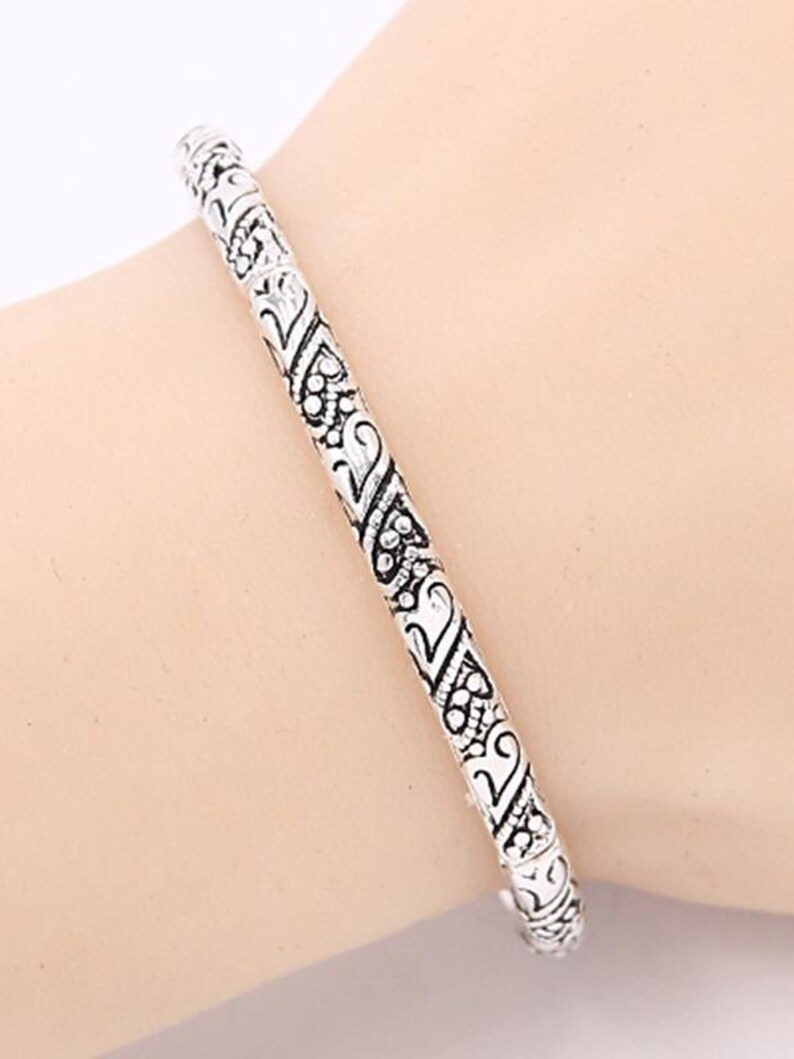May include: A silver bangle bracelet with an intricate, ornate design. The bracelet is made of metal and has a smooth, polished finish.