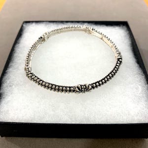 May include: A silver bracelet with a detailed design, presented in a black box with a white interior. The bracelet features a series of small, round beads and decorative elements. The overall aesthetic is elegant and detailed.