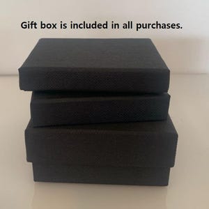 May include: Three stacked black gift boxes with textured surfaces. The boxes are rectangular and vary slightly in size, creating a tiered effect. The boxes are arranged on a white surface.