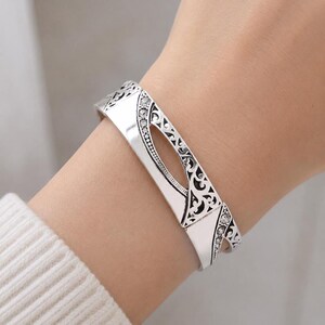 May include: A silver-toned cuff bracelet with an openwork design and clear crystal accents. The bracelet features an oval cutout in the center and decorative filigree patterns along the edges. The bracelet is worn on a wrist.