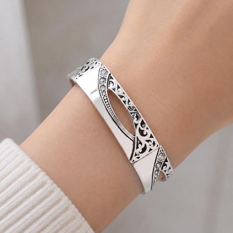 May include: A silver-toned cuff bracelet with an openwork design and clear crystal accents. The bracelet features an oval cutout in the center and decorative filigree patterns along the edges. The bracelet is worn on a wrist.