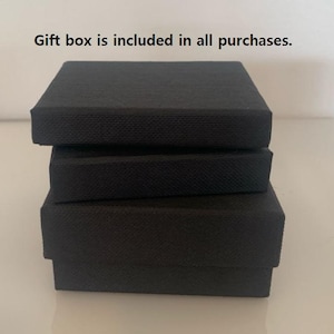 以下が含まれることがあります： テクスチャ加工された表面を持つ、3つの黒いギフトボックスが積み重ねられています。ボックスは長方形で、サイズがわずかに異なり、層状の効果を生み出しています。ボックスは白い表面に配置されています。