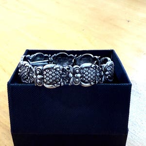 May include: A silver-toned bracelet with an ornate, textured design, resting inside a dark blue box. The bracelet features repeating floral or leaf-like patterns. The box is a simple, rectangular shape, providing a contrasting backdrop for the jewelry.