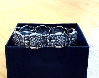 May include: A silver-toned bracelet with an ornate, textured design, resting inside a dark blue box. The bracelet features repeating floral or leaf-like patterns. The box is a simple, rectangular shape, providing a contrasting backdrop for the jewelry.