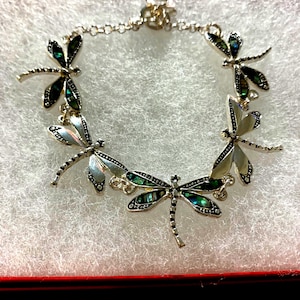 Può includere: Bracciale argentato con libellule e dettagli iridescenti verdi e neri. Il bracciale presenta sei ciondoli a forma di libellula dettagliati collegati da una delicata catena. Le ali della libellula hanno un aspetto brillante e strutturato, presentato su una superficie bianca.