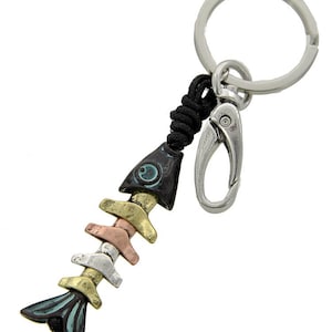 Puede incluir: Un llavero de metal con forma de esqueleto de pez, con un llavero y un mosquetón plateados. El diseño de espina de pescado presenta una mezcla de colores, incluyendo dorado, plateado, cobre y verde azulado. El llavero está sujeto a un cordón negro tejido.