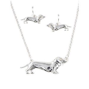 May include: A silver necklace and earrings set featuring a dachshund design. The necklace has a silver chain and a pendant of a dachshund with a single clear stone for an eye. The earrings are dangle earrings with a dachshund design.