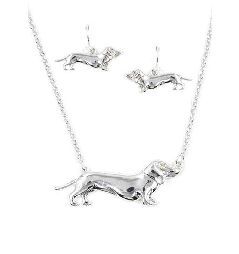May include: A silver necklace and earrings set featuring a dachshund design. The necklace has a silver chain and a pendant of a dachshund with a single clear stone for an eye. The earrings are dangle earrings with a dachshund design.