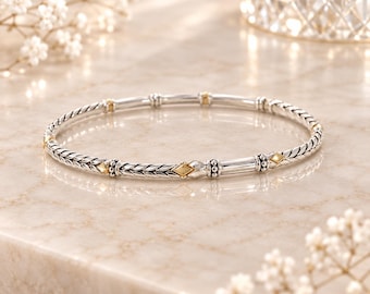 Peut inclure: Un bracelet jonc circulaire fin composé de métal argenté poli et d'accents dorés. La bande présente un motif tressé texturé alternant avec des segments cylindriques lisses et de petites incrustations en forme de losange de couleur dorée.